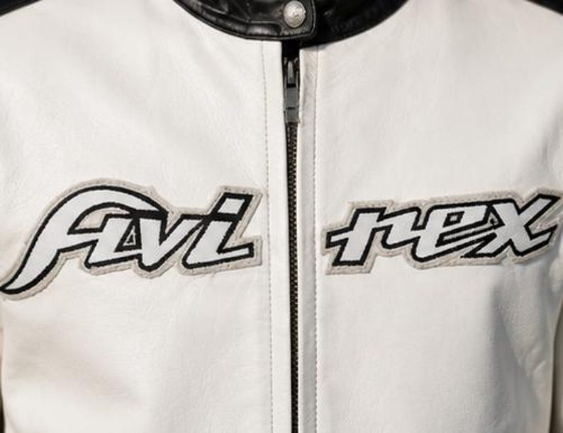 May include: Close-up of a white leather jacket with black accents. The jacket features the embroidered text "Avi rex" in black and white. A black zipper runs down the center, and the collar is black.