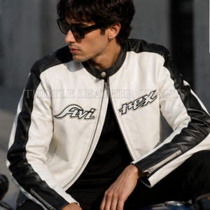 May include: A white and black leather jacket with the text "Avi Rex" on the front. The jacket has black sleeves and a black collar. The jacket is zipped up and has a classic motorcycle jacket design.