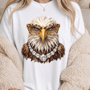 May include: A white t-shirt featuring a graphic illustration of a bald eagle wearing leopard print glasses and a necklace made of large, round, clear faceted crystals. The eagle has brown and white feathers and a yellow beak, centered on the chest.