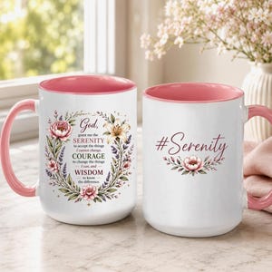 May include: Two white ceramic mugs with pink interiors and handles. One mug features a floral wreath surrounding the text God, grant me the SERENITY to accept the things I cannot change, COURAGE to change the things I can, and WISDOM to know the difference. The other mug displays the text #Serenity with a single flower.