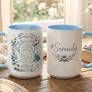 May include: Two white ceramic mugs with light blue interiors and handles. One mug features a floral wreath surrounding the Serenity Prayer text. The other mug displays the text #Serenity in blue script above a single floral illustration.