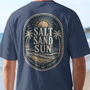May include: A navy blue t-shirt featuring a large oval graphic on the back. The design includes a sun, palm trees, and a beach scene with the text SALT SAND SUN, SIMPLE DAYS, and GOOD WAVES, along with an anchor icon in a distressed style.