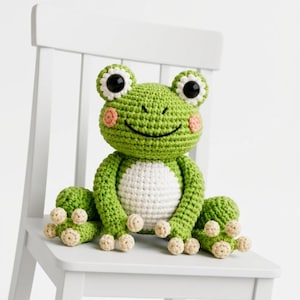 May include: A handmade green crochet frog with a white belly, sitting on a white chair. The frog has large black eyes, pink cheeks, and beige feet. The frog is a whimsical, handcrafted item.