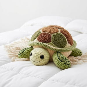 Puede incluir: Un peluche de tortuga hecho a mano con ganchillo. La tortuga tiene una cabeza y un cuerpo beige, con un caparazón en tonos marrones, verdes y beige. La tortuga tiene una cara sonriente y aletas verdes. El juguete está sobre una superficie blanca.
