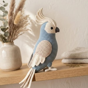Puede incluir: Un loro de ganchillo hecho a mano en tonos azules, beige y gris. El ave tiene una cresta, alas y plumas de cola distintivas. Se exhibe en una estantería de madera, mostrando la artesanía del estilo amigurumi. Un elemento decorativo.