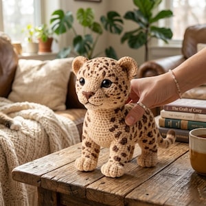 May include: A crocheted leopard cub in tan and brown. The handmade plush animal has dark brown spots and large, dark eyes. It is standing on a wooden table. The background includes a couch, plants, and books.