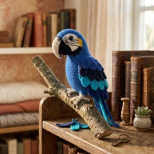 Puede incluir: Un loro azul tejido a mano con un pico crema y detalles negros, posado en una rama de madera. El ave hecha a mano se exhibe en una estantería de madera, junto a libros antiguos, lo que sugiere un objeto decorativo.