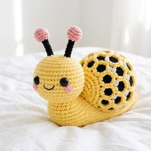Puede incluir: Un juguete de caracol hecho a mano con ganchillo en amarillo, negro, rosa y blanco. El caracol tiene un cuerpo amarillo, una concha con un patrón de panal de abeja negro y blanco y antenas rosas. El caracol tiene una cara sonriente con ojos negros y mejillas rosas.