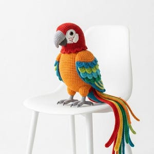 Puede incluir: Un loro de ganchillo hecho a mano en colores vibrantes. El ave presenta una cabeza roja, cuerpo naranja y alas y cola multicolores. Está posado en una silla blanca, mostrando los intrincados detalles del trabajo con hilo.