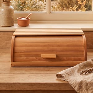 May include: A wooden bread box with a curved top and a handle sits on a wooden countertop. A small glass bottle of oil, a sprig of rosemary, and a linen cloth are on the right. A vase of wheat and lavender sits near a window.