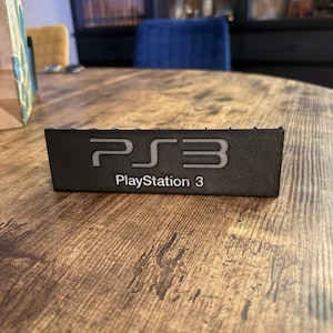 May include: A black rectangular PlayStation 3 console, with the PS3 logo and "PlayStation 3" text in silver. The console is on a wooden table.