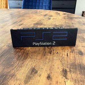 May include: A black PlayStation 2 console with blue accents and the words "PS2" and "PlayStation 2" printed on the front. The console is rectangular and sits on a wooden table.