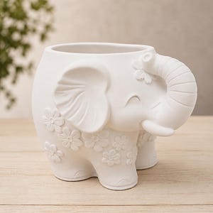 Può includere: Fioriera a forma di elefante in ceramica bianca con un volto sorridente e dettagli floreali. La proboscide dell'elefante è arricciata verso l'alto e i fiori sono posti attorno al corpo. La fioriera è progettata per contenere piccole piante o succulente.