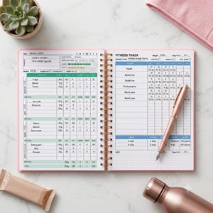 May include: A pink spiral-bound fitness planner open to a page with a meal plan and fitness track. The left page lists daily nutrition, while the right page tracks exercises, sets, reps, and weights in lbs. A rose gold pen rests on the right page.