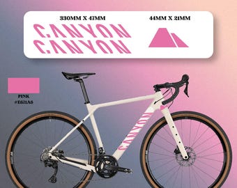 Könnte beinhalten: Ein Set aus rosa Fahrradrahmenaufklebern mit dem Canyon-Logo und einer Berggrafik. Die Aufkleber messen 330mm x 47mm und 44mm x 21mm. Die Farbe ist Pink mit dem Hex-Code #E671A6. Die Aufkleber werden über einem cremefarbenen Rennrad dargestellt.