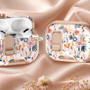 May include: Two floral-patterned AirPod cases, one open with white earbuds inside. The cases are gold-colored with a clear overlay featuring a colorful floral design. One case has a silver keychain attached. The background is a peach-colored satin.