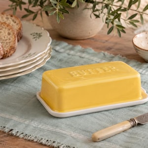 May include: A bright yellow ceramic butter dish with a rectangular lid featuring the word BUTTER embossed on the top. The dish sits on a white base and is accompanied by a silver-toned knife with a cream-coloured handle, placed on a light green cloth.