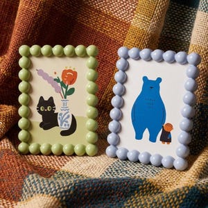 May include: Two decorative picture frames. One frame is light green with a black cat illustration and a vase of flowers. The other frame is light blue with a blue bear and a small person illustration. Both frames have a beaded border.