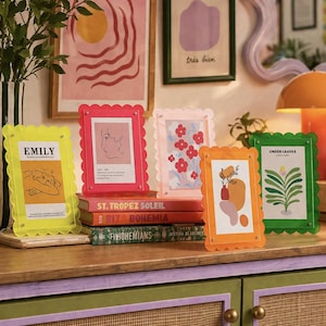 Könnte beinhalten: Eine Sammlung farbenfroher Acryl-Bilderrahmen mit gewellten Kanten. Jeder Rahmen enthält einen anderen Kunstdruck, darunter Blumendesigns, abstrakte Formen und Text. Die Rahmen sind auf einer Holzoberfläche mit Büchern angeordnet.