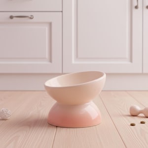 May include: A ceramic pet bowl with a two-tone design. The top is a light cream color, and the base transitions from a light pink to a deeper coral. The bowl has a tilted, oval shape and a rounded base. A small white ball and a toy are in the background.