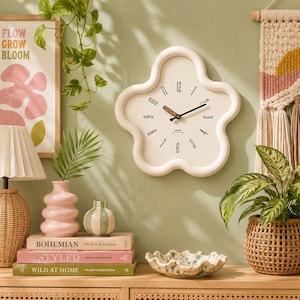 Puede incluir: Un reloj de pared crema en forma de nube con manecillas negras y números minimalistas. El reloj está montado en una pared verde claro, complementado con elementos decorativos como jarrones, libros y una cesta tejida. También es visible una impresión enmarcada con las palabras "FLOW GROW BLOOM".