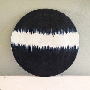 Op de afbeelding: Een ronde, zwarte decoratieve plaat met een brede, horizontale witte band. De witte band is versierd met verticale, donkerblauwe penseelstreken, waardoor een gestructureerd, artistiek ontwerp ontstaat. De plaat is waarschijnlijk voor decoratieve doeleinden.
