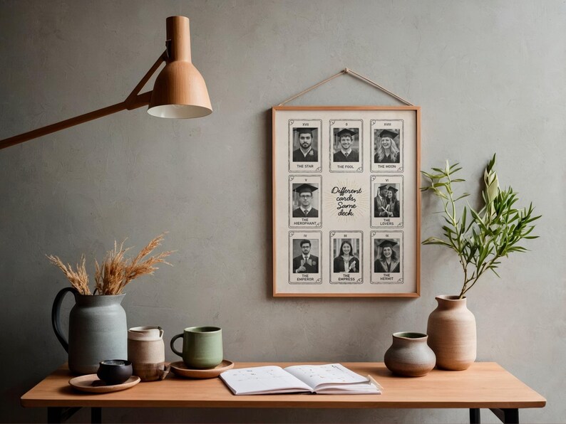 May include: A framed print displaying nine black and white tarot-style cards with graduation photos. The text "Different cards, Same deck" is centered on the print. The print is suspended on a grey wall above a wooden table with vases, mugs, and an open book.