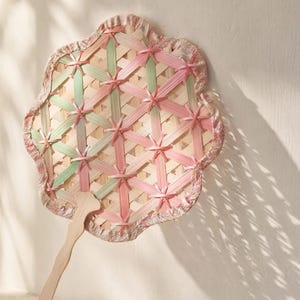 May include: A hand fan featuring a woven bamboo lattice structure with alternating light pink and pale green strips. The fan has a scalloped edge finished with a floral patterned fabric trim and a light-coloured wooden handle attached at the base.