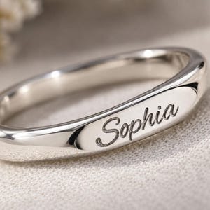 Puede incluir: Un anillo de banda en tono plateado pulido con un perfil liso y redondeado y una sección rectangular plana en el centro. El nombre Sophia está grabado en una fuente cursiva oscura sobre la superficie plana del anillo de metal.
