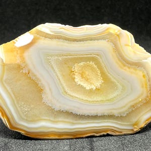 May include: A polished agate stone slice with concentric bands of cream, white, and golden-yellow hues. The center features a lighter, oval-shaped pattern. The stone's edges are a darker yellow, creating a natural, decorative piece.