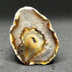 Può includere: Una fetta di agata lucida con un motivo naturale a strati. La pietra presenta fasce di tonalità bianche, marroni e gialle, con una cavità centrale. I bordi dell'agata sono bianco crema e la forma generale è irregolare.