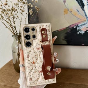 May include: Cream-colored phone case with a quilted pattern featuring butterflies and floral designs. The case has a brown leather strap and gold-colored hardware. The phone case has multiple camera lenses.
