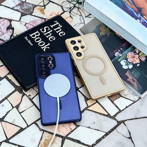 May include: Two phone cases, one blue and one gold, with MagSafe chargers attached, rest on a patterned surface. A black book titled "The She Book" is also present. The cases are designed for a smartphone.