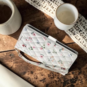 Puede incluir: Funda de teléfono acolchada blanca con un estampado floral en rosa, rojo y verde. La funda tiene una correa para la muñeca y un cierre magnético. El fondo incluye una superficie de madera, una taza blanca y un elemento decorativo blanco tejido.