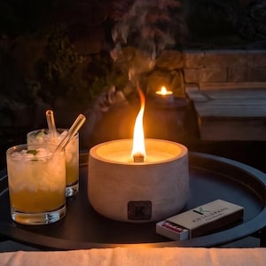 Könnte beinhalten: Eine runde, hellgraue Beton-Feuerschale mit einer hellen orangefarbenen Flamme, zwei Gläser mit Eis und einem gelben Getränk sowie eine kleine rechteckige Schachtel mit der Aufschrift KIESELBEANS auf einem schwarzen runden Tablett.
