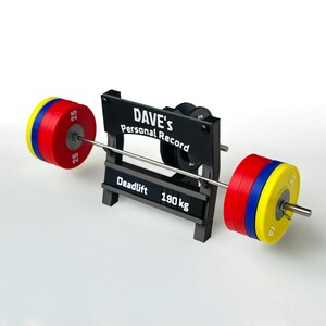 Könnte beinhalten: Ein schwarzer Hantelständer mit der Aufschrift DAVE'S Personal Record und Deadlift 190 kg. Der Ständer hält eine Miniatur-Langhantel mit roten, blauen und gelben Gewichtsscheiben auf jeder Seite, wobei die Zahl 25 auf den roten und 15 auf den gelben Scheiben zu sehen ist.