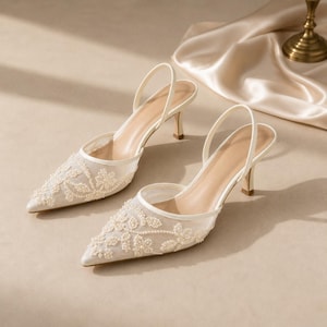 May include: A pair of cream-colored slingback heels featuring a pointed toe and a slim kitten heel. The front of the shoes is crafted from sheer mesh fabric adorned with intricate floral embroidery and small white beads in a matching tone.