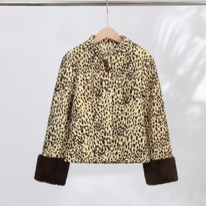 May include: A long-sleeve jacket featuring a cream and dark brown leopard print pattern. The jacket has a round neckline, a front button closure with three dark brown buttons, and wide, solid dark brown faux fur cuffs at the end of each sleeve.