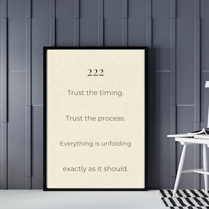 May include: A rectangular wall art print in a black frame featuring a cream-colored background with the black text: 222, Trust the timing., Trust the process., Everything is unfolding, exactly as it should. The design is centered and minimalist.