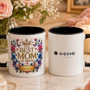 Puede incluir: Dos tazas de cerámica blancas con asas y bordes negros. Una taza presenta diseños florales y el texto "The Best Mom is Mine" con el nombre "Dayana". La otra taza tiene un código escaneable y el texto "Scan to Listen".