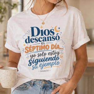 Puede incluir: Camiseta blanca con el texto "Dios descansó el séptimo día... yo solo estoy siguiendo su ejemplo" en azul y naranja. La camiseta tiene las mangas dobladas y se combina con unos vaqueros azules. El texto es de Génesis 2:2.