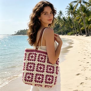 Può includere: Una borsa tote all'uncinetto con un motivo ripetuto di granny square nelle tonalità magenta, rosa scuro e crema. La borsa ha una forma rettangolare con base piatta e due tracolle in pelle marrone fissate con elementi in metallo.