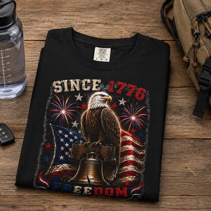 May include: A black t-shirt featuring a graphic print of a bald eagle perched on a bell, with an American flag, fireworks, and the text "SINCE 1776" and "FREEDOM" in red, white, and blue colors. The shirt has a size L-G-G label at the neck.