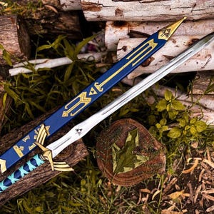 K&ouml;nnte beinhalten: Ein silbernfarbenes Schwert mit blau-goldfarbenem Griff und einer passenden blauen Scheide mit goldenen Akzenten. Das Schwert liegt auf einem Bett aus Holz und Gr&uuml;n, das eine Waffe im Fantasy-Stil zeigt.