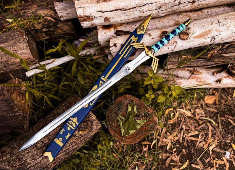 K&ouml;nnte beinhalten: Ein silbernes und goldenes Fantasy-Schwert mit einer blauen und goldenen Scheide. Das Schwert hat einen detaillierten Knauf und eine Klinge. Die Scheide ist mit goldenen Akzenten und Symbolen verziert. Das Schwert und die Scheide liegen auf Holzst&auml;mmen und Gr&uuml;n.