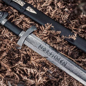 May include: A close-up of a Viking sword with a black handle and a silver blade. The blade is engraved with the word "NORTHMEN". The sword is lying on dried leaves, with a black scabbard visible in the background.