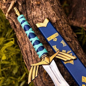 K&ouml;nnte beinhalten: Ein dekoratives Schwert mit goldenem Knauf und silberner Klinge, das auf einer Holzoberfl&auml;che liegt. Der Griff ist mit blauen und t&uuml;rkisfarbenen B&auml;ndern umwickelt. Eine blaue Scheide mit goldenen Akzenten liegt daneben.