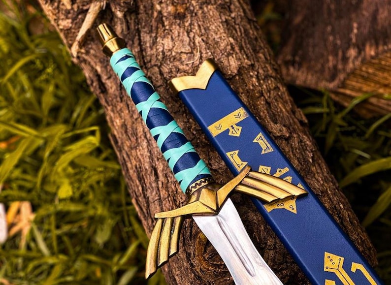 K&ouml;nnte beinhalten: Ein dekoratives Schwert mit goldenem Knauf und silberner Klinge, das auf einer Holzoberfl&auml;che liegt. Der Griff ist mit blauen und t&uuml;rkisfarbenen B&auml;ndern umwickelt. Eine blaue Scheide mit goldenen Akzenten liegt daneben.
