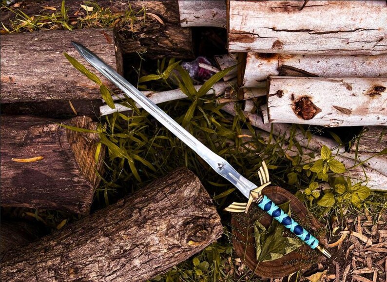 K&ouml;nnte beinhalten: Ein Silberschwert mit blau-goldenem Griff liegt auf einem Bett aus Baumst&auml;mmen und Gr&uuml;n. Das Schwert hat eine lange, spitze Klinge und einen dekorativen Knauf mit goldenen Akzenten. Der Griff ist mit blauem Leder mit Rautenmuster umwickelt.