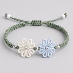 May include: A macrame bracelet featuring two flower-shaped charms, one cream and one light blue, with silver beads. The bracelet is adjustable with a green cord and white beads at the ends.